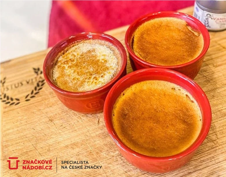 Tři krémy brûlée s karamelizovanou vrstvou v červených miskách na dřevěném podkladu.