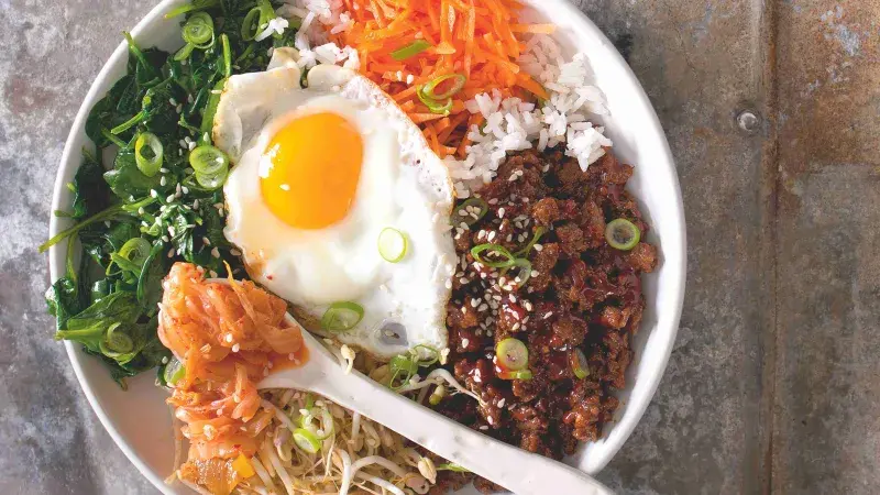 Bibimbap s vejcem, zeleninou a křupavým masem na bílém talíři.