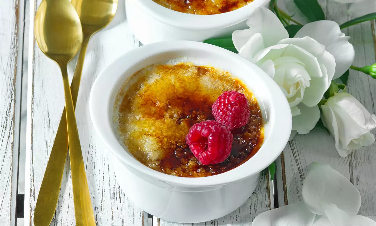 Klasická crème brûlée s malinami v bílém hrnci na dřevěném stole.