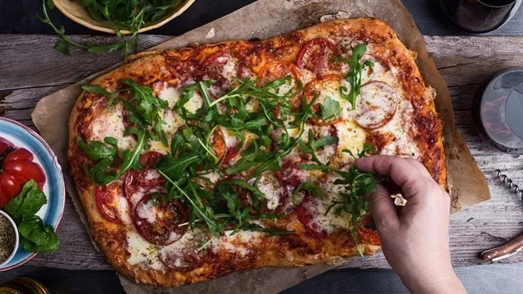 Ruce přidávají rukolu na domácí pizzu s rajčaty.
