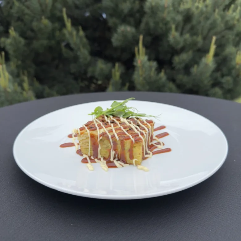 Kousek okonomiyaki s omáčkou na bílém talíři venku.
