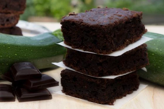 Hromada čokoládových brownies vedle cuket na dřevěném povrchu.