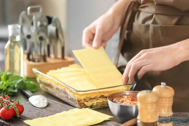 Príprava lasagne s vrstvami těstovin, rajčat a zeleniny na dřevěném stole.