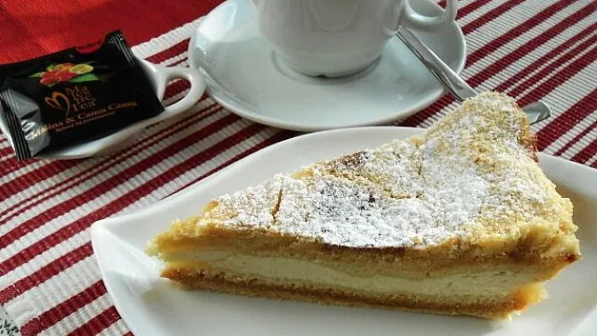 Kousek tvarohového koláče posypaného cukrem na bílém talíři.