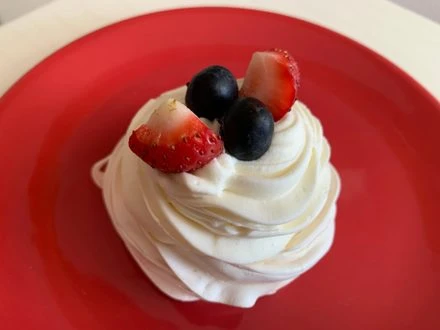 Bílá merengová Pavlova s jahodami a borůvkami na červeném talíři.