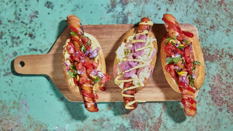 Tři domácí hot dogy s omáčkami na dřevěném podnosu.
