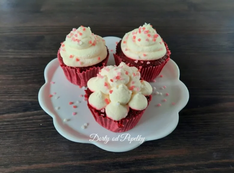 Tři červené muffiny s bílým krémem na šlehačku a cukrovými strouhankami.