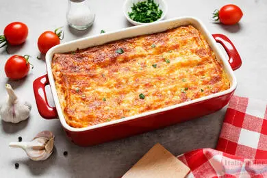 Pečená lasagne v červené misce s čerstvými ingrediencemi kolem.