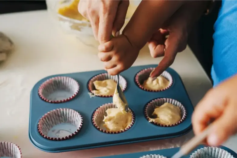 Ruce nalévají těsto do papírových formiček na muffiny.