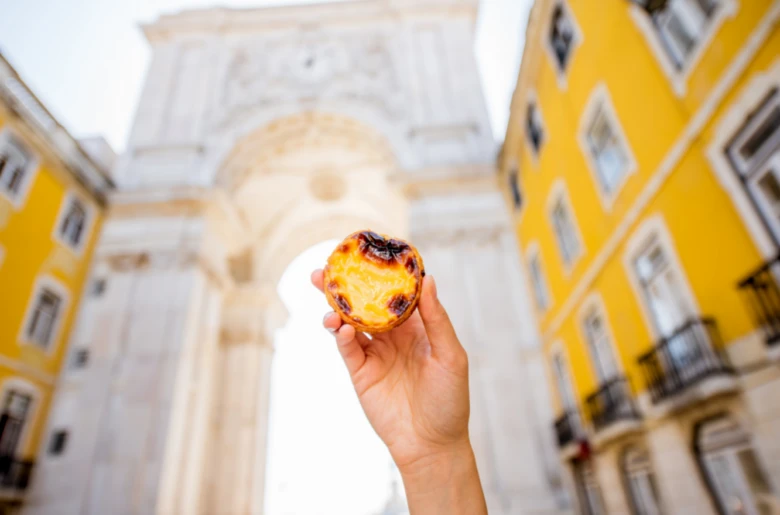 Ruka drží portugalský dezert pastel de nata před historickou bránou.
