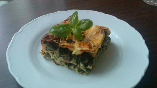 Kousek pečené lasagne se špenátem na bílém talíři s bazalkou.