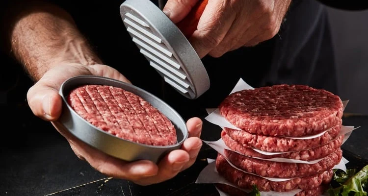 Ruce tvarují syrové hovězí burgery pomocí lisu vedle hotových plack.