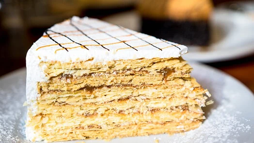 Kousek dortu napoleon s vrstvami krému na bílém talíři.