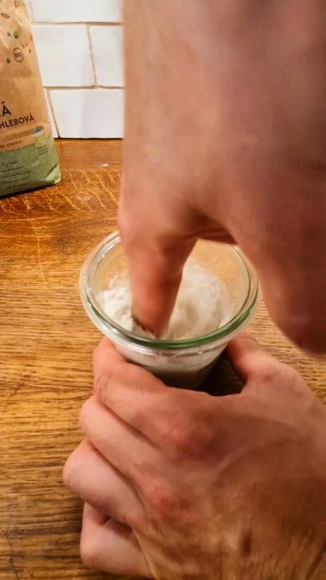 Ruka míchá mouku v malém skleněném kelímku na dřevěné desce.