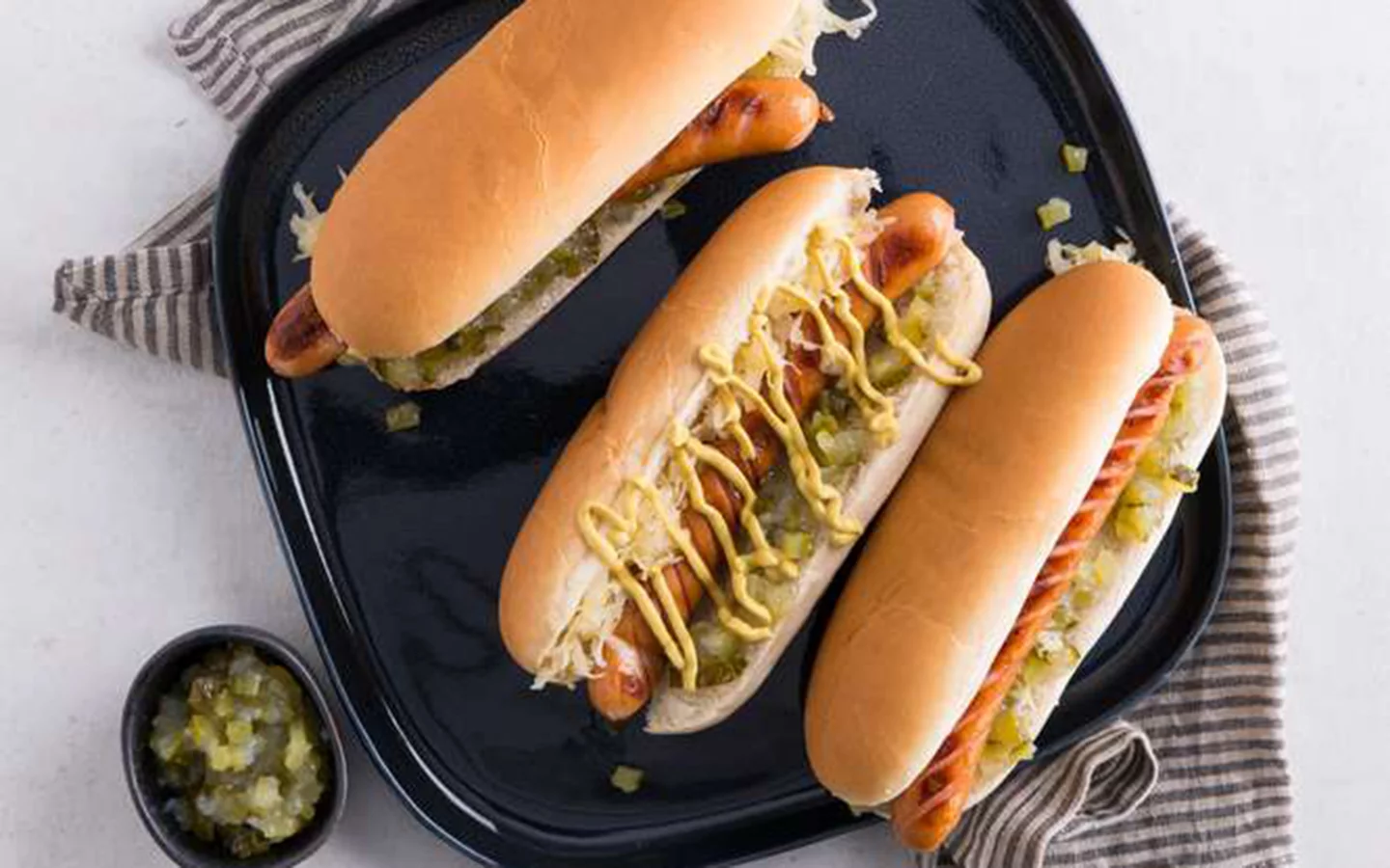 Tři domácí hot dogy s hořčicí na černém podnosu.