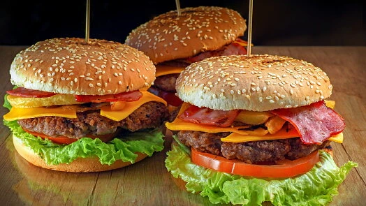 Tři domácí burgery s kuřecími masy, sýrem a slaninou na dřevěném povrchu.