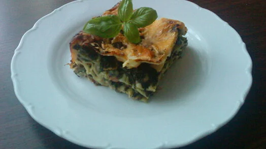 Kousek pečené lasagne s mletým masem, špenátem a bazalkou na bílém talíři.