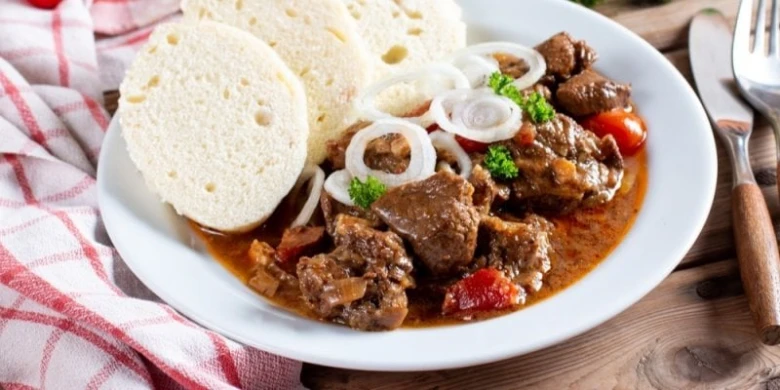 Český guláš s knedlíky a cibulovými kroužky na talíři.